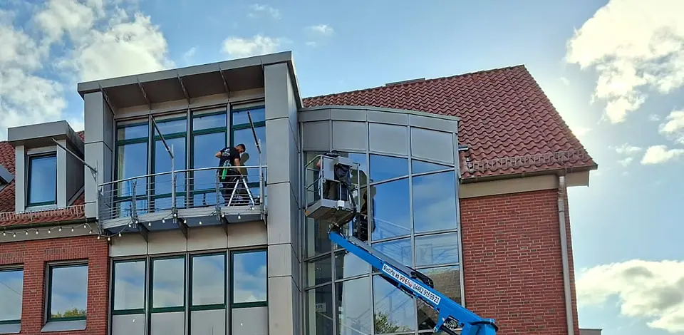 Glasfassade eines Geschäftshauses: ein Mitarbeiter reinigt vom Balkon aus, einer von der Arbeitsbühne